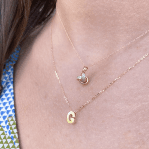 Two gold necklaces on a woman's chest. The top neckle is a love know. The bottom necklace is a letter "G" pendant.