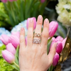 womans hand with two 3-row bezel set diamond rings stacked on middle finger in front of pink tulips and blue hydrangeas