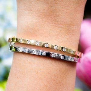 close up view of womans hand and wrist infront of blue and pink flowers wearing two bezel set diamond station bracelets