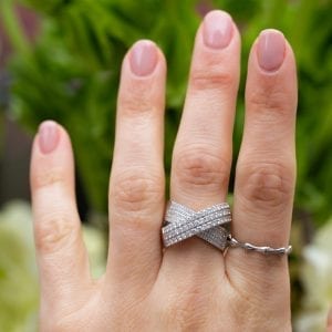 white gold and diamond crossover bridge ring on womans middle finger infront of greenery